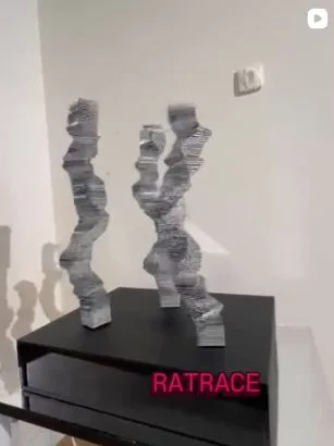 Ratrace — twee gestapelde sculpturen op een zwarte sokkel met neonletters
